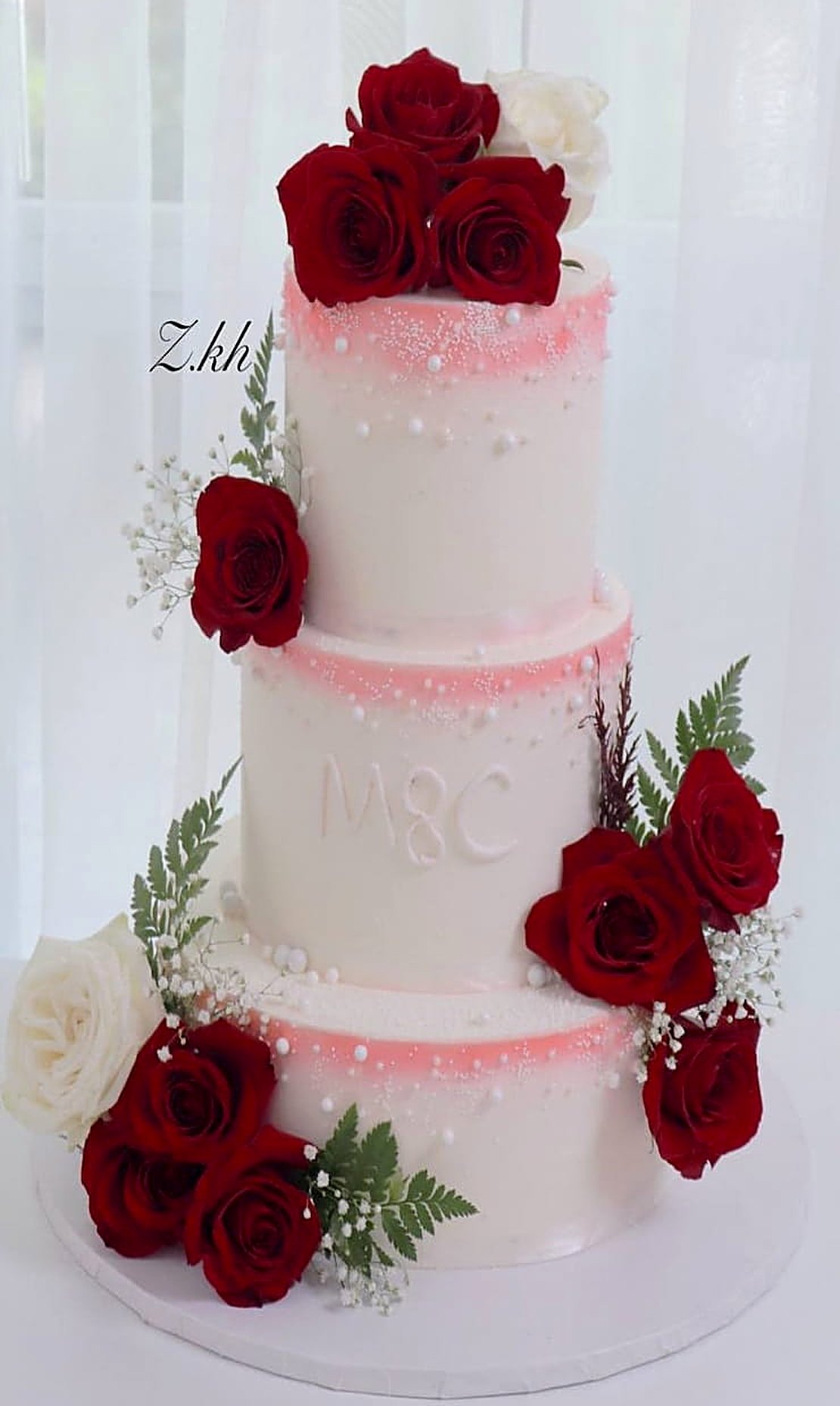 Three-tier blush and white wedding cake with red roses, initials M & C